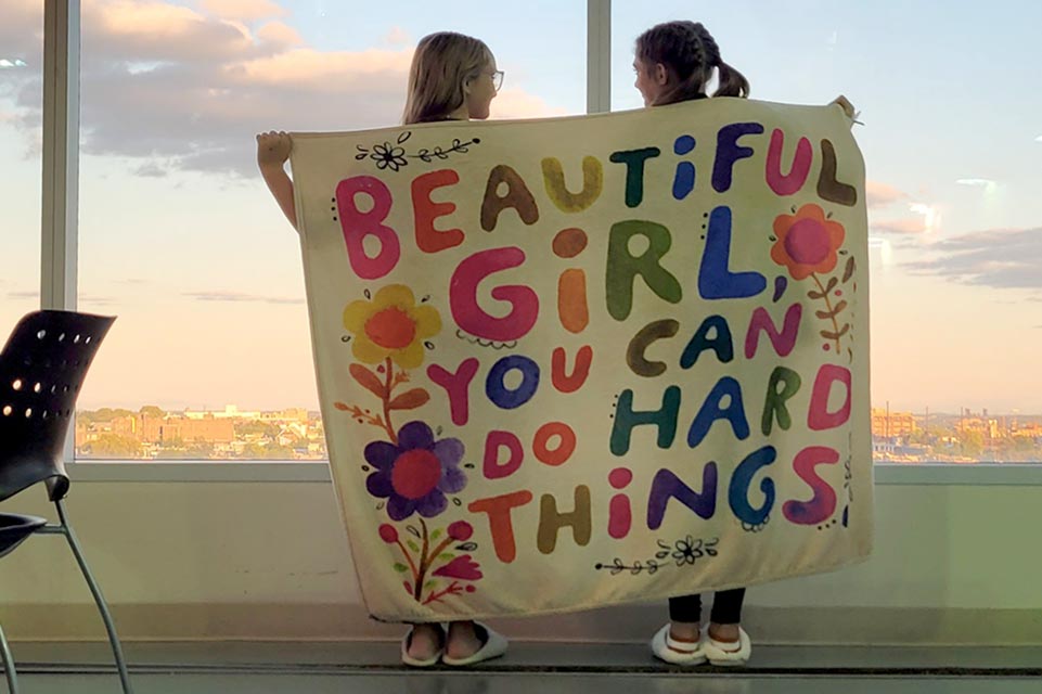 Macie y Lou Lou sostienen una pancarta que dice Beautiful Girl, You can Do Hard Things (Bella niña, puedes lograr grandes cosas)