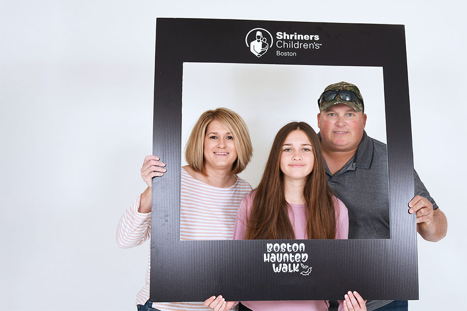 Female patient with her parents holding a frame with Shriners Children's Boston logo and Boston Haunted Walk logo