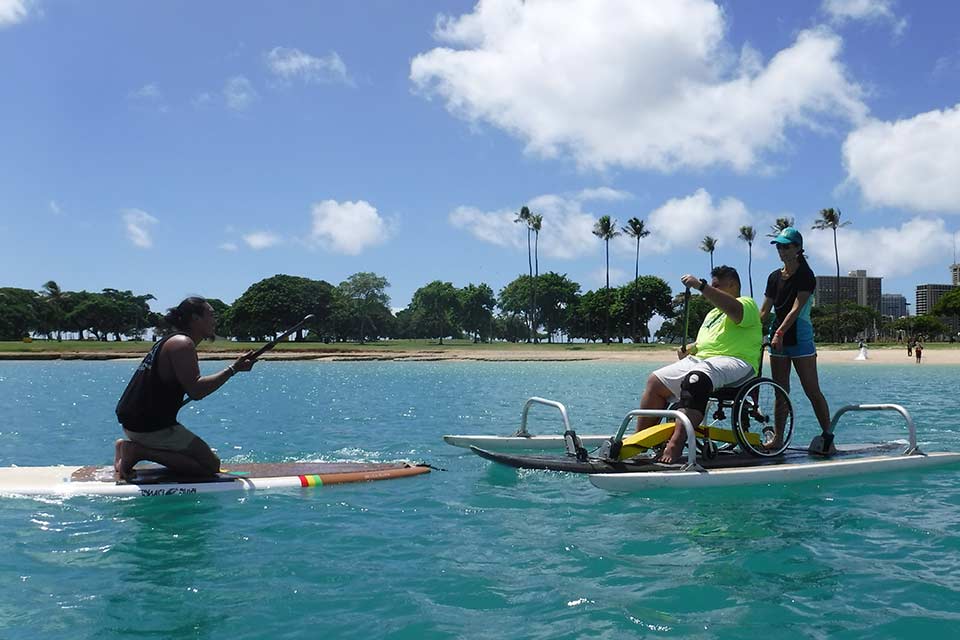 Un joven varón en una tabla surf de remo adaptada con una ayudante mujer.